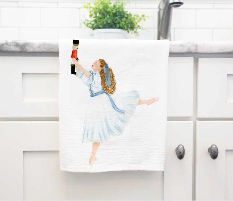 A white cotton tea towel featuring a watercolor illustration of a Clara from the Nutcracker in a blue dress, holding a Nutcracker. The setting is in a kitchen environment.