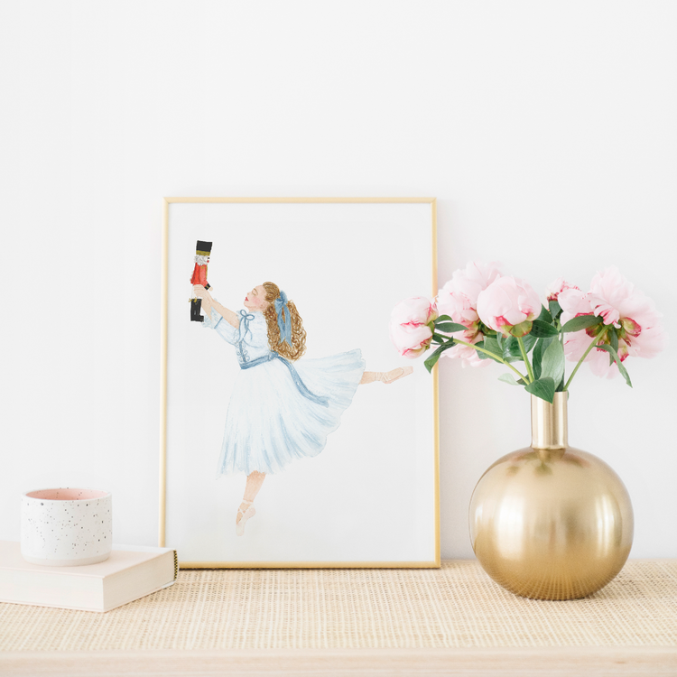An art print featuring Clara from the Nutcracker in a white dress and holding a Nutcracker doll. There is a gold vase with pink flowers in the photo.