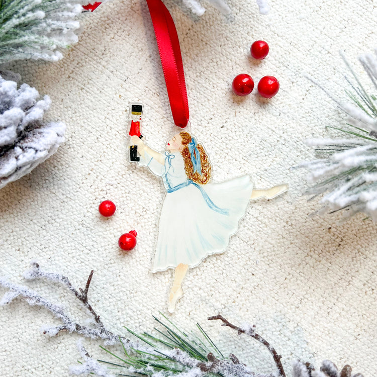 An ornament featuring Clara from the Nutcracker holding a Nutcracker doll with a red ribbon at the top of the ornament. The background has red berries and faux greenery