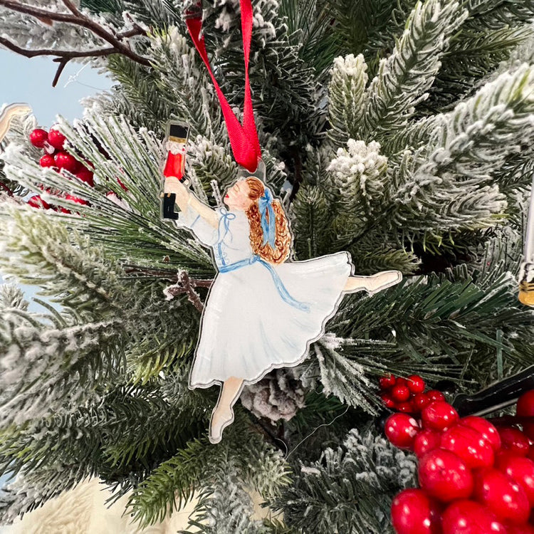 An ornament hanging on a tree with a red ribbon featuring Clara from the Nutcracker. The tree also has red berries.