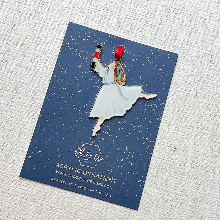 An ornament featuring Clara from the Nutcracker wtih a red ribbon on a navy blue backer card.