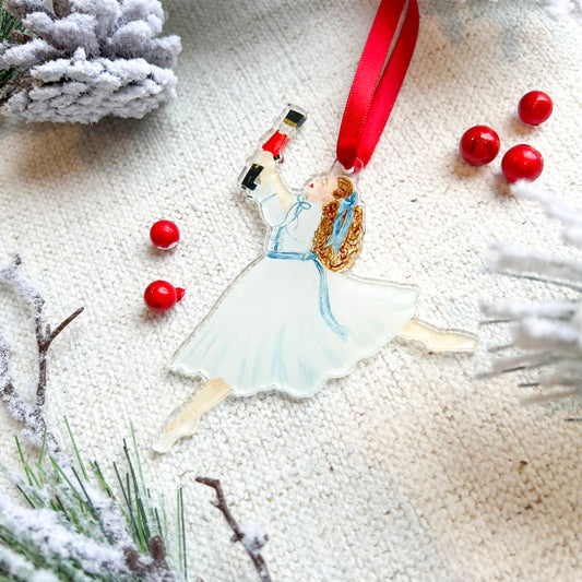 A holiday ornament featuring a watercolored illustration of Clara from the Nutcracker, with red ribbon for hanging.