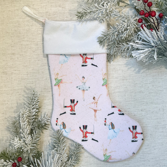 Handmade pink velvet Nutcracker ballet stocking with a white cuff, decorated with images of ballerinas and nutcrackers, displayed against a Christmas-themed backdrop.