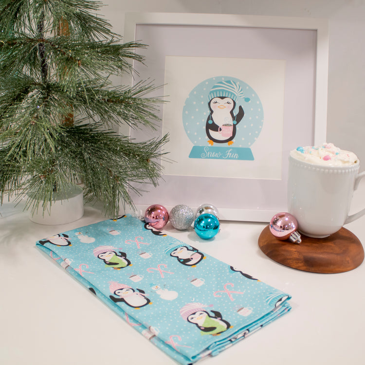 A blue and white winter penguin-themed tea towel displayed on a wooden holder, with a framed picture of a penguin in the background and Christmas decorations.