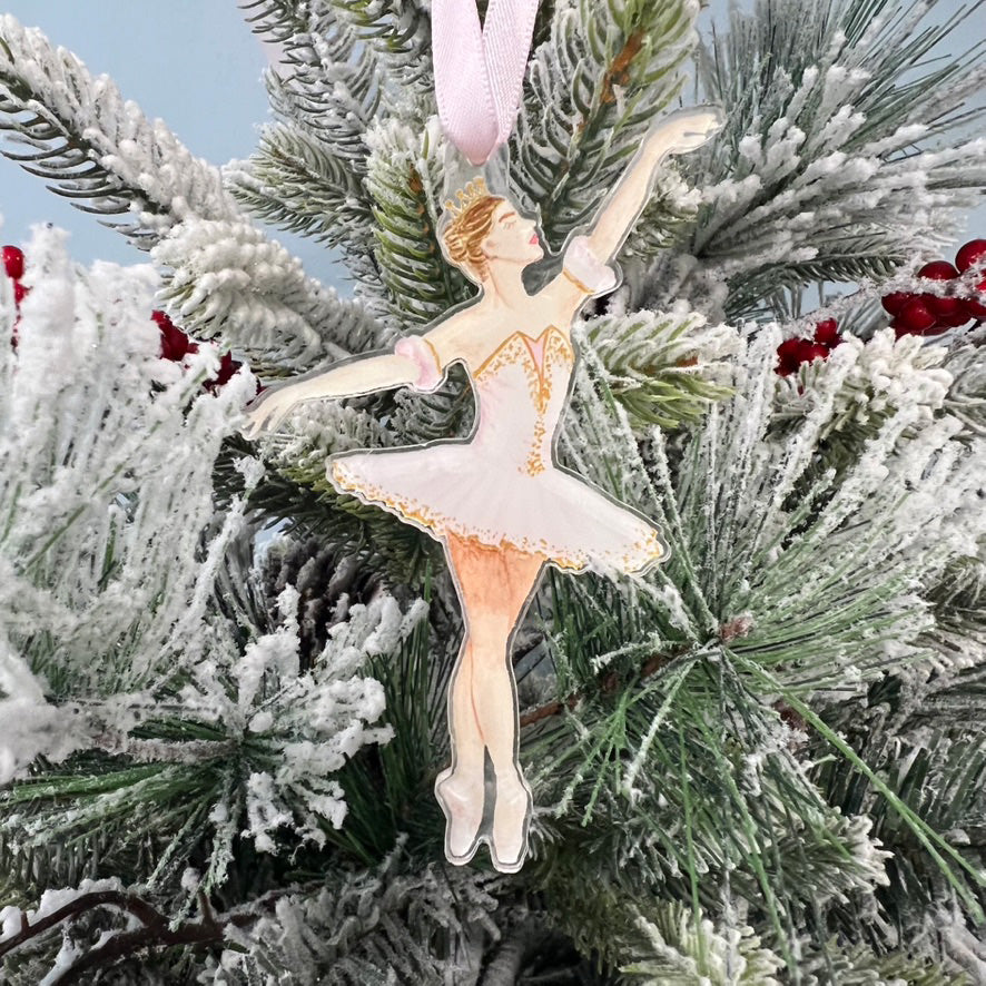 An ornament featuring Sugarplum Fairy from the Nutcracker wearing a pink tutu. The background has a branches from a Christmas tree.