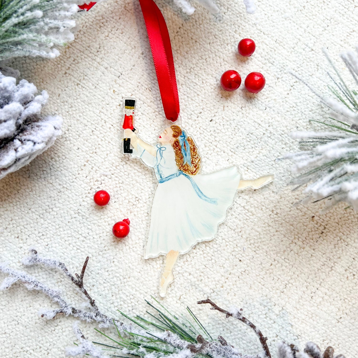 An ornament featuring Clara from the Nutcracker holding a Nutcracker doll with a red ribbon at the top of the ornament. The background has red berries and faux greenery