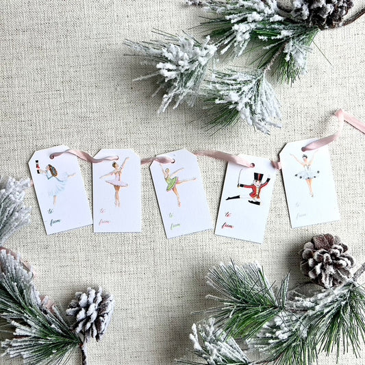 A set of white gift tags with watercolor illustrations of Nutcracker themed characters, including a nutcracker and a snow queen, interspersed with pink ribbon. The tags are displayed against a backdrop of green pine needles and snowflakes.
