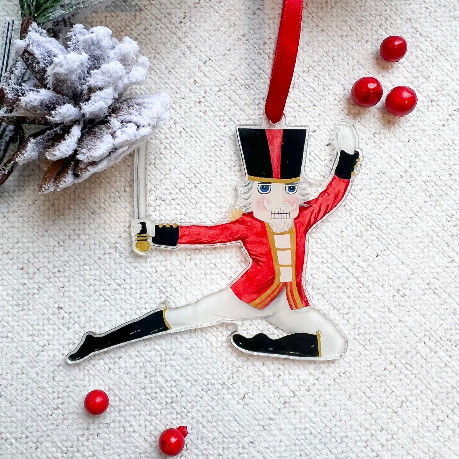 Nutcracker Prince ornament hanging on a red ribbon with red berries and a pinecone in the background.